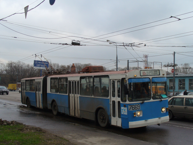 Ein blauer und weißer Oberleitungsbus mit vielen Passagieren fährt an einem Auto auf einer Straße vorbei, die mit Gras und Steinen gesäumt ist, mit Strommasten, Bäumen, Gebäuden und einem bewölkten Himmel im Hintergrund.