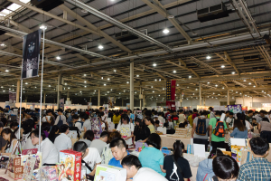 Eine große Gruppe von Menschen umringt Tische mit Büchern, Kartons und anderen Gegenständen auf einer Buchmesse, mit Schildern und Lichtern darüber.