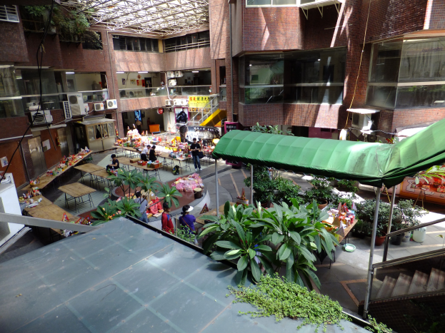 Großer Atrium mit üppiger Vegetation, Menschen, Tischen, Stühlen, Treppen mit Geländern, Gebäuden mit Fenstern und Klimaanlagen sowie einem Schuppen im Hintergrund.