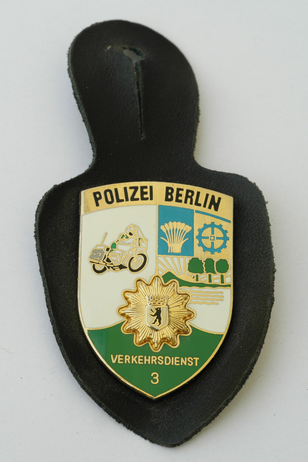 Ein Berliner Polizeistern mit blauem Schild, weißer Umrandung, zentralem weißen Stern und schwarzer 'Polizei Berlin'-Aufschrift in fetter Schrift, liegt auf einer Oberfläche.
