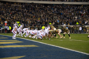 Ein Fussballspiel im Gange mit Spielern in Sportbekleidung und Helmen auf dem Feld, Zuschauern, Geländern und Tafeln mit Text im Hintergrund.