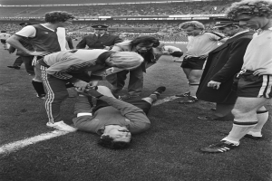 Schwarzes und weißes Bild eines Fußballspielers, der auf dem Boden liegt und von einer Gruppe Männer umgeben ist, mit einer Zuschauermenge im Hintergrund eines Stadions.