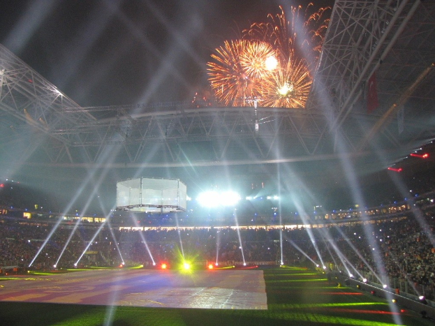 Olympiastadion in London bei Nacht, erleuchtet von hellen Lichtern, mit einer großen Menge Menschen, grünem Rasenfeld und Feuerwerk, das den Himmel erhellt.