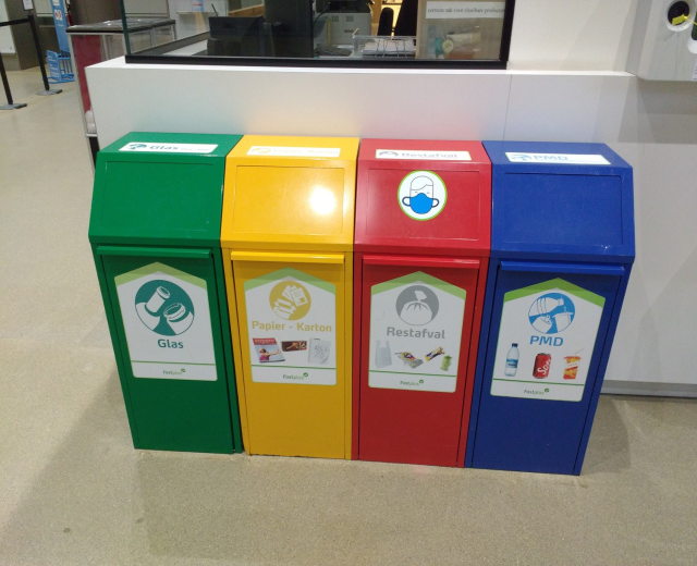 Vier bunte Mülltonnen in einer Reihe auf dem Boden mit einer Theke, Monitor und anderen Gegenständen im Hintergrund, die wahrscheinlich Recyclingbehälter in Singapur darstellen.