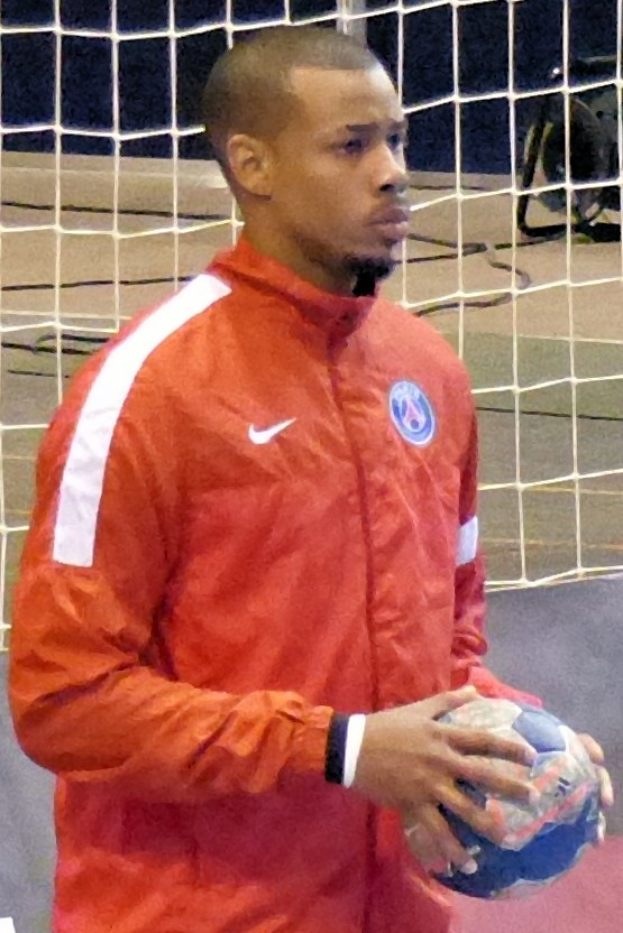 Ein Mann in einem blauen T-Shirt und Shorts steht vor einem Fußballtor und hält einen Fußball mit entschlossener Miene, kurzes dunkles Haar und fokussierte Augen.