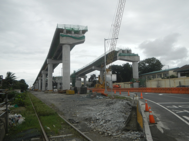Baustelle mit einer Brücke im Hintergrund, Straße durch Verkehrskegel markiert, Bahnschiene links, verstreute Steine und Gras, Bäume und Gebäude auf beiden Seiten und ein bewölkter Himmel.
