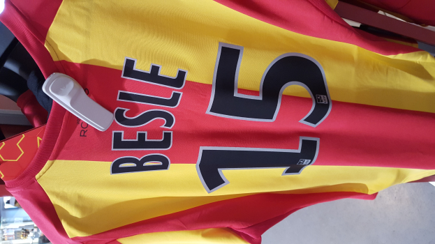 Roter und gelber Fußballtrikot mit aufgedrucktem Text und Nummern an einer Kleiderstange hängend, mit anderen Gegenständen und dem Boden im Hintergrund.