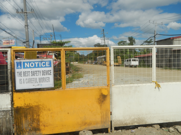 Ein gelbes Tor mit einem Sicherheitshinweisschild, dahinter sind Strommasten, Bäume, Schuppen, Fahrzeuge auf einer Straße und ein bewölkter Himmel.