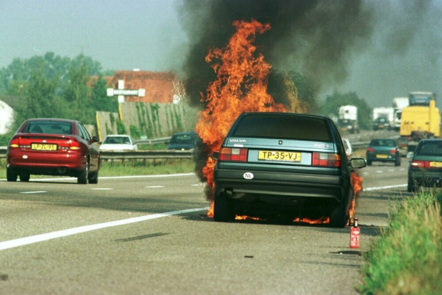 Ein brennendes Auto am Straßenrand mit umliegenden Fahrzeugen, Bäumen, Gebäuden und einem klaren blauen Himmel im Hintergrund und einem Feuerlöscher auf der rechten Seite.