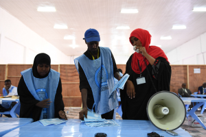 Eine Gruppe von Menschen steht um einen Tisch mit einem Megafon, Papieren und anderen Gegenständen herum, die scheinbar eine Wahllokalität darstellen, mit anderen im Hintergrund auf Stühlen sitzend und stehend unter beleuchteter Decke.