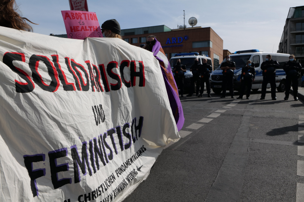 Gruppe von Menschen, die eine 'Solidarität und Feminismus'-Schlagzeile tragen, marschieren auf einer Straße mit geparkten Fahrzeugen, Gebäuden, einer Schüsselantenne und einem klaren blauen Himmel im Hintergrund.