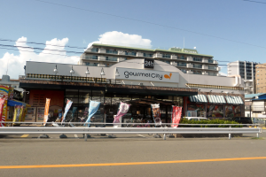 Eine belebte Stadtstraße mit Gebäuden, Straßenmöbeln, Fahrzeugen, Grünflächen und einem Restaurant, das Speisen in seinem Fenster präsentiert.
