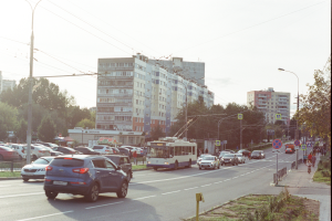 Eine belebte Stadtstraße mit zahlreichen Autos, Bussen, Straßenlaternen, Schildern, Lichtmasten, Strommasten mit Drähten, Bäumen und Gebäuden unter einem klaren blauen Himmel.