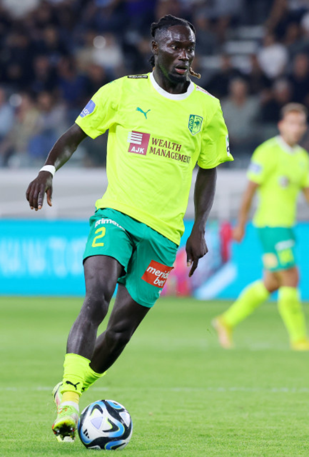 Ein Fußballspieler in gelbem Trikot und grünen Shorts ist im Einsatz auf dem Feld, mit einem blauen Board und unscharfen Figuren im Hintergrund.