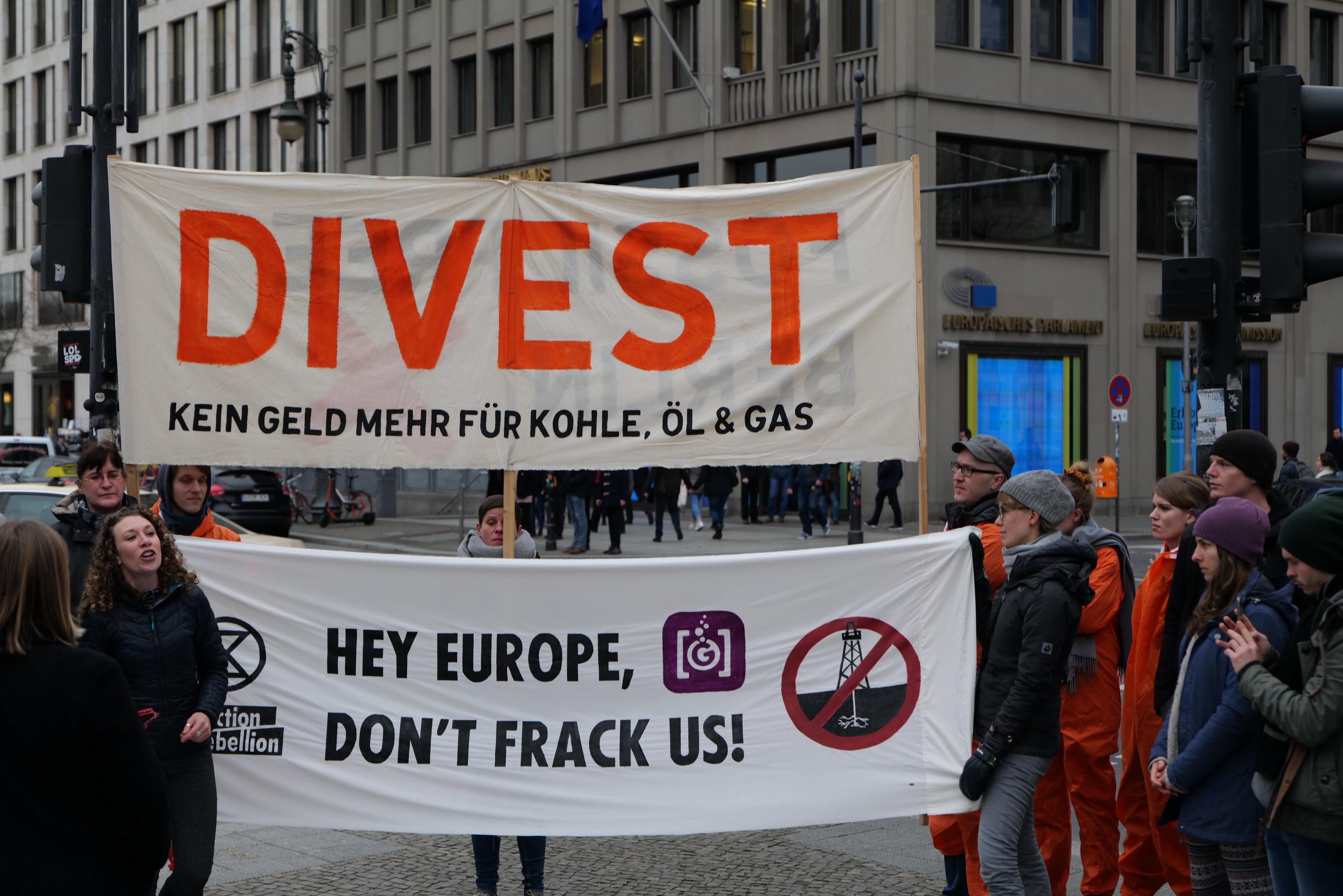 Gruppe von Menschen mit einem Banner 'Divest Hey Europe, Don't Frack Us' auf einer Straße mit Gebäuden, Straßeninfrastruktur, Fahrzeugen, Bäumen und einem klaren Himmel im Hintergrund.