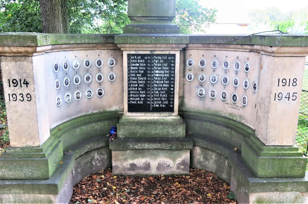 Ein Holocaust-Gedenkstein in einem jüdischen Friedhof in Berlin, mit einer Wand voller Text und Zahlen, umgeben von Bäumen und einem Zaun mit verstreuten trockenen Blättern.