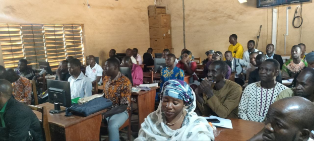 Eine Gruppe von Menschen sitzt an Tischen in einem Klassenzimmer mit Monitoren, Taschen, Büchern und Papieren und nimmt an einem Techniktraining teil; links ist ein Fenster zu sehen, und es gibt Kartons und einen Fernseher an der Wand im Hintergrund.