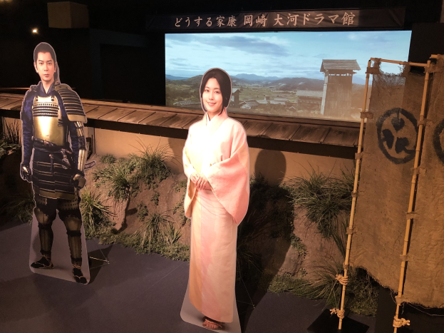 Zwei Personen - eine Frau in einem rosa Kimono und ein Mann in Samurai-Rüstung - stehen lachend in einer traditionellen japanischen Umgebung mit Pflanzen, einem Bildschirm und Beleuchtungsausrüstung.