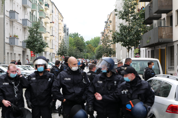 Eine Gruppe von Polizisten in Masken und Helmen, die eine von geparkten Autos gesäumte Straße entlanggehen, flankiert von Gebäuden mit Balkonen und Geländern, mit Bäumen und einem klaren Himmel im Hintergrund.