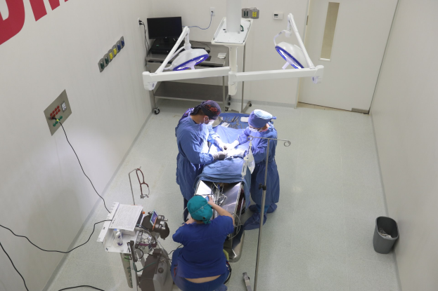 Chirurgen führen eine Operation an einem Patienten auf einer Trage in einem Operationssaal durch, mit medizinischer Ausrüstung an einer Wand und einem Objekt auf dem Boden rechts.