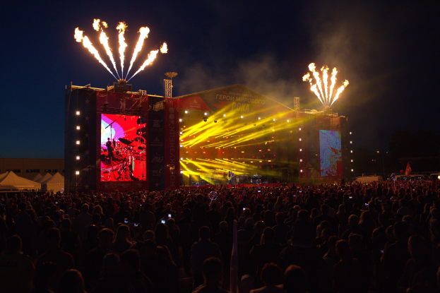 Eine große Menschenmenge bei einem Musikfestival schaut auf einer hell erleuchteten Bühne mit Bannern, umgeben von Zelten und Feuerwerk am dunklen Himmel.