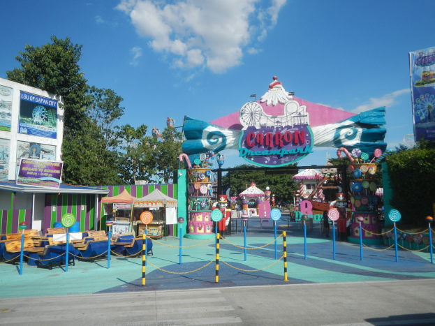 Karussell am Parkeingang mit Straße darunter, eingezäuntes Gebiet, Buden, Banner, Bäume und bewölkter Himmel im Hintergrund.