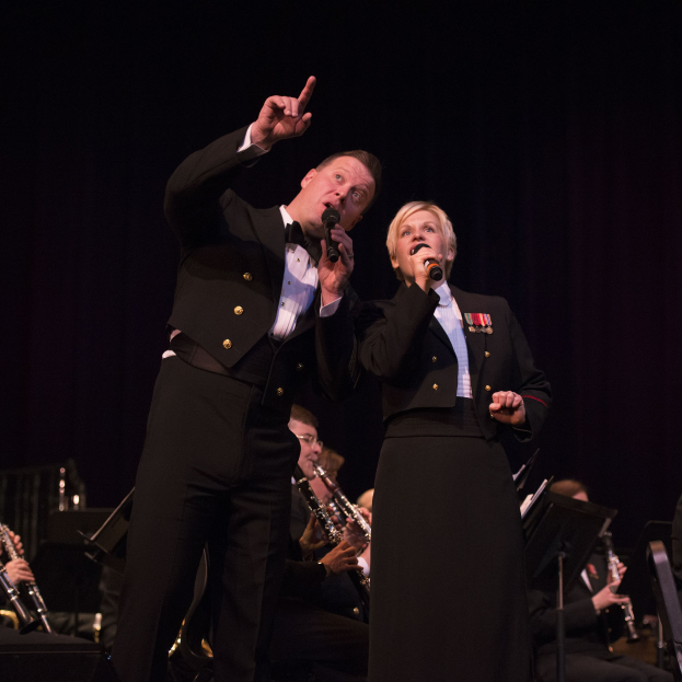 Ein Mann und eine Frau in Fracks, die Mikrofone auf einer Bühne halten, mit einem sitzenden Orchester dahinter vor einem dunklen Hintergrund.