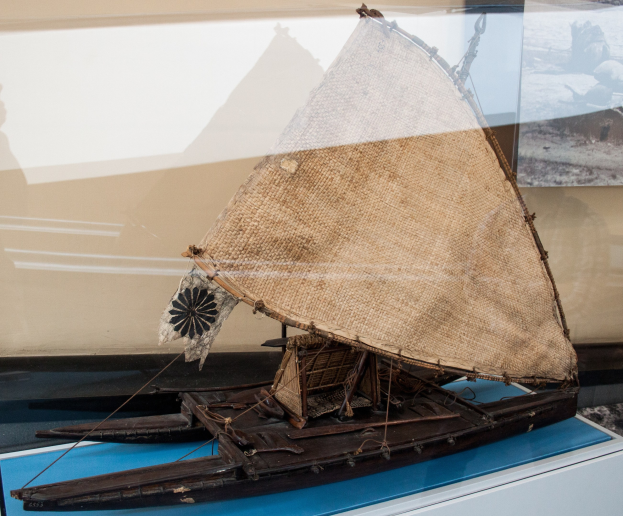 Ein hölzernes Modellboot mit einem Segel in einer Glasvitrine, mit einem Foto Rahmen im Hintergrund.