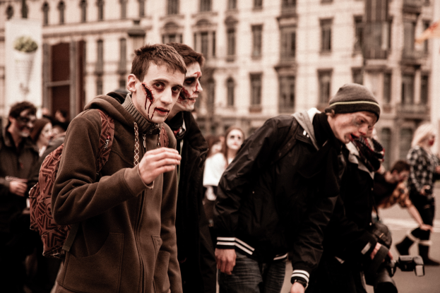 Eine Gruppe von Menschen in Zombie-Kostümen geht eine Straße entlang, wobei einige Kostüme tragen und einer eine Kamera hält, und ein Gebäude im Hintergrund zu sehen ist.