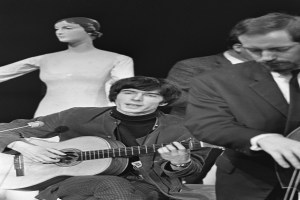 Ein Mann in einem schwarzen und weißen Outfit sitzt auf einem Stuhl und spielt eine Gitarre neben einer Schaufensterpuppe, mit einem Mikrofon vor ihnen, vor einem dunklen Hintergrund, als Hommage an den verstorbenen Paul McCartney.