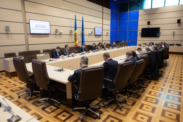 Große Gruppe von Menschen an einem langen Tisch mit Mikrofonen und Papieren in einer formellen Besprechungssituation, mit Flaggen, Fernsehern und Deckenleuchten im Hintergrund.