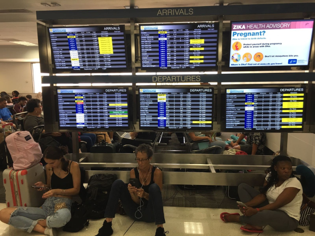 Eine Gruppe von Menschen sitzt auf dem Boden eines Flughafens, einige halten Handys, mit verstreuten Taschen und Bildschirmen, die Zika-Virus-Informationen anzeigen.