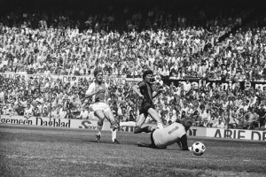 Schwarzes Foto von Männern beim Fussballspielen auf einem Feld mit Zuschauern im Hintergrund und einer "Bundesliga 1978-79 Ajax 1-1 Hannover 96"-Fahne.
