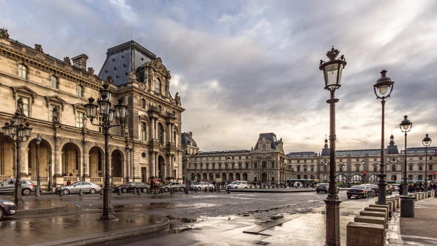Das ikonische Louvre-Museum in Paris, Frankreich, mit seinen historischen Gebäuden, der Straßeninfrastruktur, fahrenden Fahrzeugen, Passanten und bewölktem Himmel.