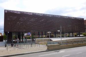 Großes modernes Gebäude mit zahlreichen Fenstern, das als neuer Eingang zur Stadtmitte dient, umgeben von Straßeninfrastruktur, Fahrzeugen, Fußgängern, Bäumen und einem bewölkten Himmel.