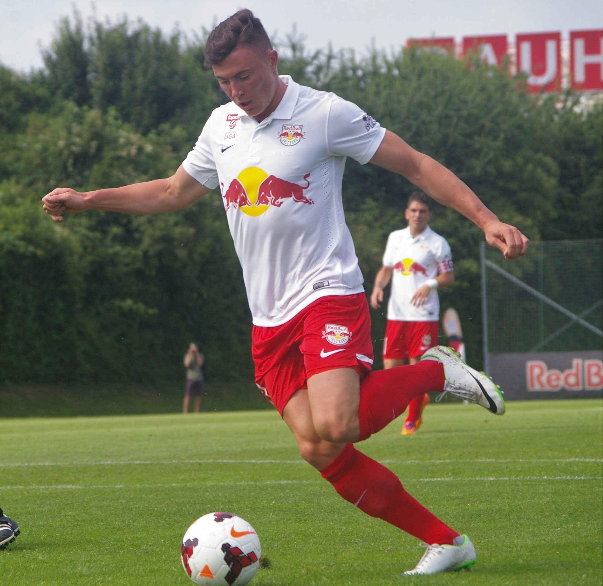 Ein professioneller Fußballspieler kickt einen Ball auf einem grünen Feld mit Bäumen und einem klaren blauen Himmel im Hintergrund, in der Nähe einer Tafel, auf der "RB Leipzig" steht.