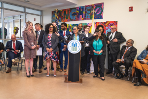 Gruppe von Menschen vor einem Podium mit Mikrofon und einer Tafel mit dem Text "Sancho City Councilman Joins City Council", mit sitzenden und stehenden Teilnehmern, Flaggen, Feuerlöscher und Deckenleuchten im Hintergrund.