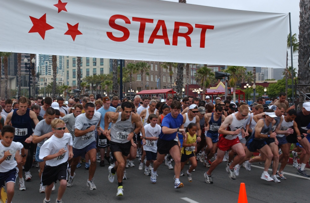 Gruppe von Läufern bei einem Marathon, die an einer Verkehrskegel vorbeilaufen, mit einem Banner und Gebäuden im Hintergrund unter einem klaren blauen Himmel.
