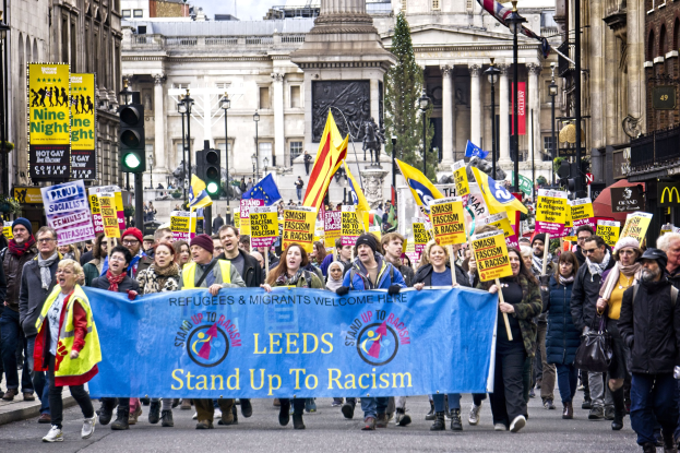 Gruppe von Menschen marschiert durch eine Stadtstraße und hölt eine 'Steh Auf gegen Rassismus'-Plakette, mit Gebäuden, Bäumen, Straßenlaternen, Ampeln und einer Statue im Hintergrund bei bewölktem Himmel.