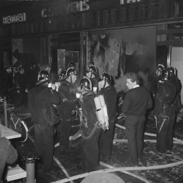 Schwarzes Bild von Feuerwehrleuten in Uniform mit Sauerstoffflaschen vor einem Gebäude, einem Fahrrad und einem Tisch mit Gegenständen.