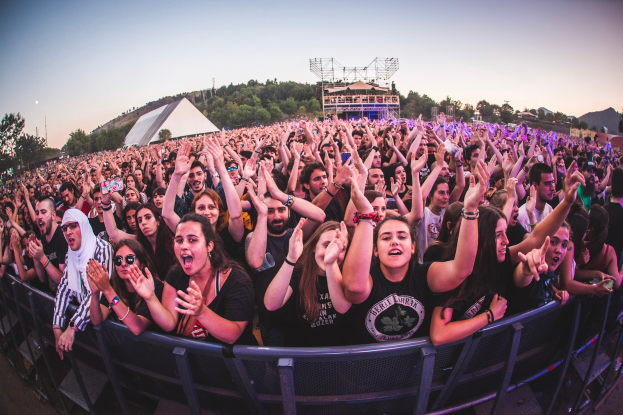 Eine große Menschenmenge auf einem Musikfestival mit erhobenen Händen vor einem Geländer mit Bäumen, Gebäuden und einem klaren blauen Himmel im Hintergrund.