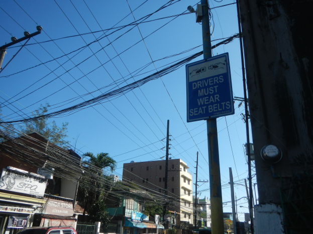 Stadtstraße mit fahrenden Autos, Strommäste mit Drähten, Gebäude, Bäume und Schilden, mit einem "Fahrer müssen den Sicherheitsgurt anlegen"-Schild an einem Strommast vor einem sichtbaren Himmel.