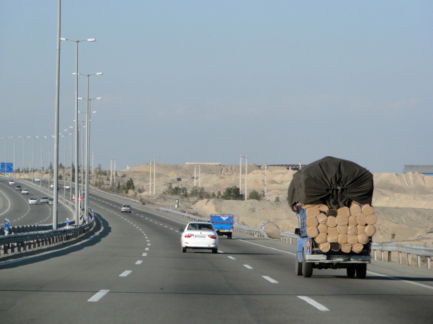 Ein Holzlastwagen fährt auf einer Straße mit einer großen Ladung Holz auf dem Anhänger, umgeben von Geländern, Laternen, Schildern, Bäumen und Sand, mit Hügeln und einem klaren blauen Himmel im Hintergrund.