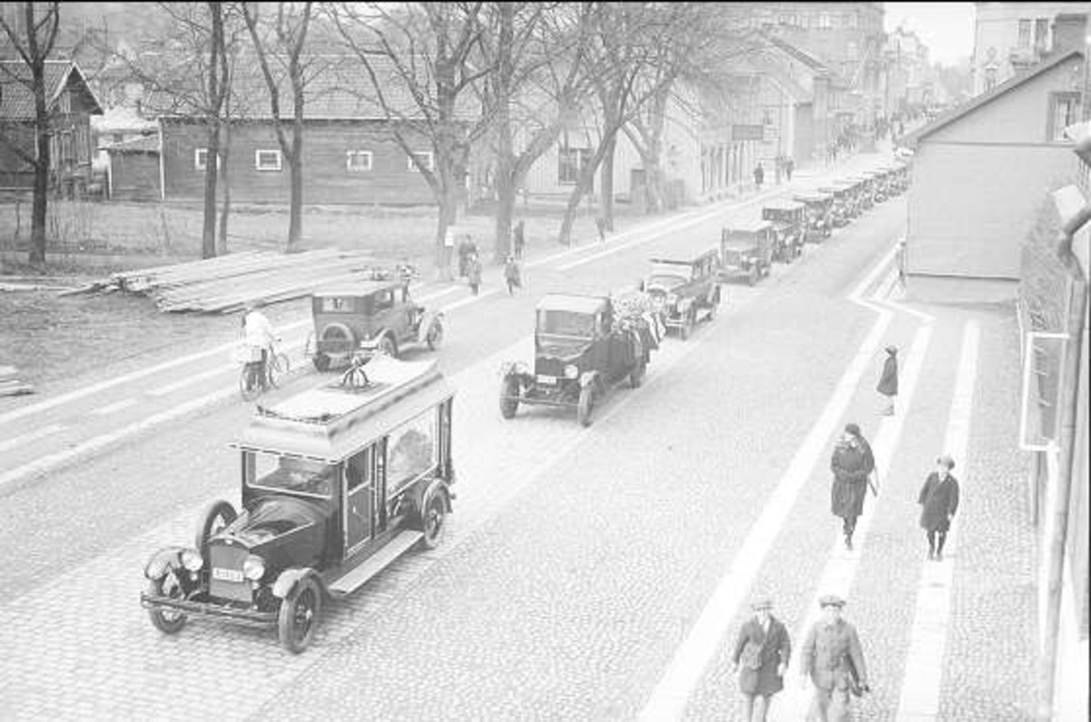 Schwarz-weißes Straßenbild in Berlin aus den 1930er-1940er Jahren, das alte Autos, Fußgänger, Gebäude mit Fenstern, Bäume und einen klaren Himmel zeigt.