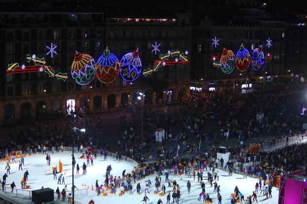 Eine große Menge von Menschen, die auf einer vor einem Gebäude beleuchteten Eisbahn Schlittschuh laufen, die mit festlichen Weihnachtsdekorationen geschmückt ist, mit Geländern und Pfosten umgeben und einem dunklen Hintergrund.
