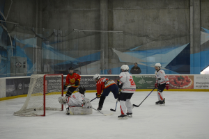 Gruppe von Menschen, die Eis Hockey auf einem Eisplatz spielen, mit Helmen und Hockeystöcken, mit einem Torpfosten auf der linken Seite, Bannern im Hintergrund und einer Wand.