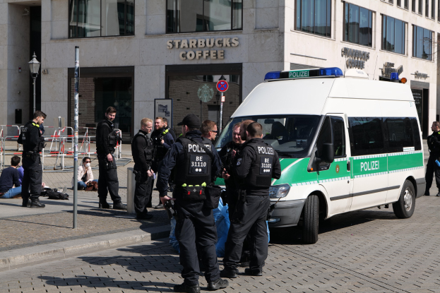 Eine Gruppe von Polizisten steht vor einem Starbucks-Café, rechts ein Van und links ein paar Menschen; im Hintergrund ist ein Gebäude mit Fenstern, ein Schild, ein Laternenpfahl und ein Zaun zu sehen.