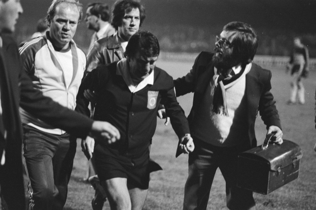 Schwarzes und weißes Bild einer Gruppe von Männern, die auf einem Fußballfeld gehen, einer hält eine Aktentasche, der zentrale Mann sieht besorgt aus.
