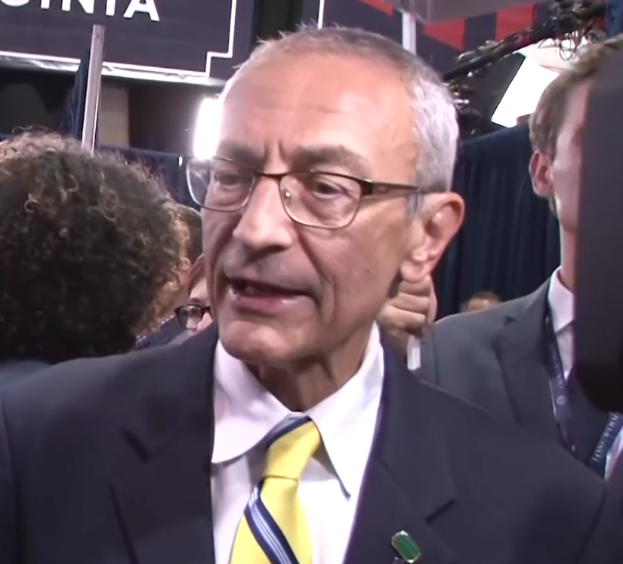 Ein Mann im Anzug und Krawatte, der eine Brille trägt, spricht mit Reportern, während ein Mikrofonständer vor ihm steht und er von einer Gruppe Menschen umgeben ist, mit Lichtern und einer Tafel im Hintergrund.