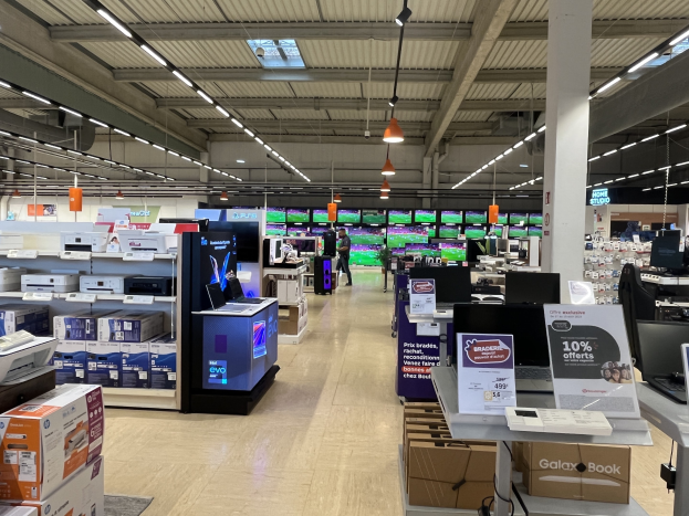 Ein großer Raum in einem Walmart-Laden voller Elektronik wie Computern und Druckern, die in Regalen angeordnet sind, mit Menschen auf dem Boden, einer Säule, Schildern, Deckenlampen und Deckenventilatoren.